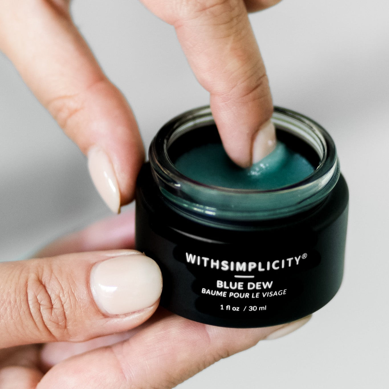 Hand holding a black jar of 'Blue Dew' with 'WITHSIMPLICITY' branding on a light background