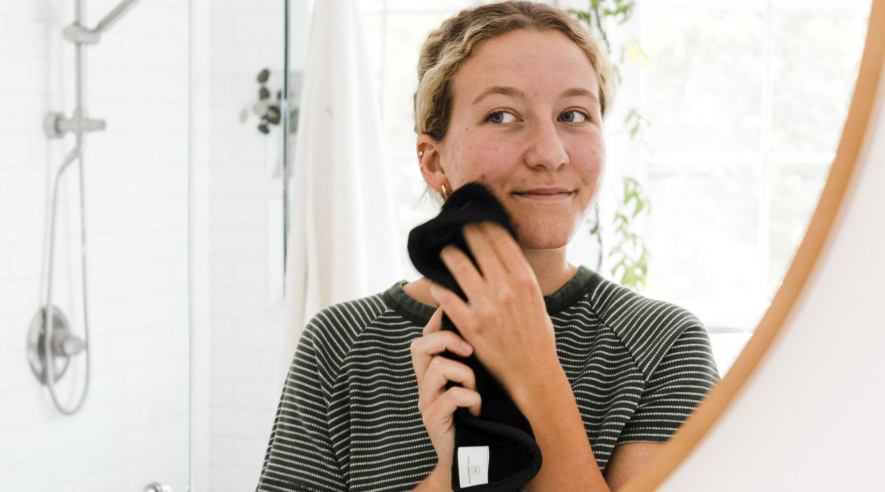 Girl holding black face wash up to face while looking into a mirror in a bathroom setting