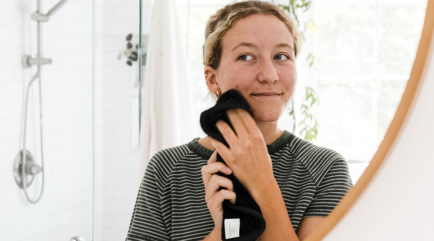 Girl holding black face wash up to face while looking into a mirror in a bathroom setting