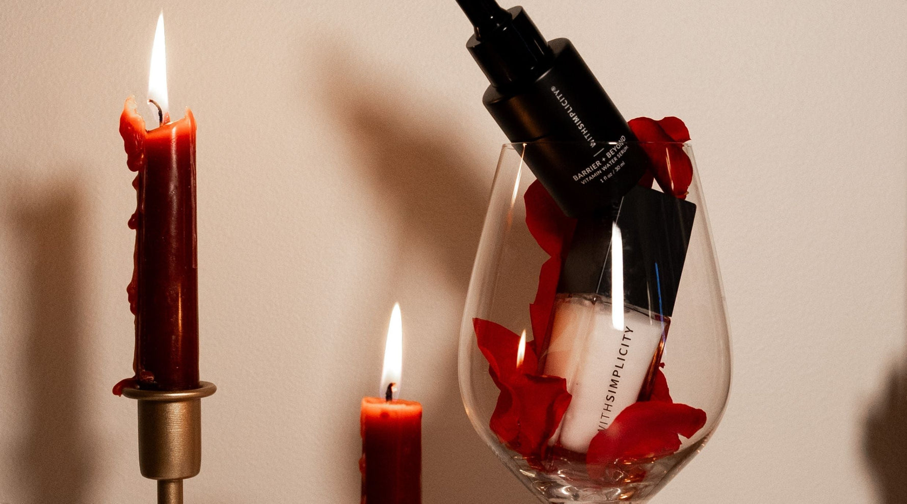A romantic close-up of withSimplicity’s Barrier + Beyond Vitamin Water Serum and Face Primer resting inside a clear wine glass filled with red rose petals next to a red candle with mood lighting and a tan background