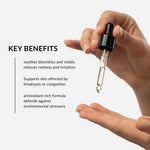 A hand holds a small cosmetic dropper bottle above another hand, with text on the left reading 'KEY BENEFITS' and bullet points about soothing blemishes, reducing redness and irritation, supporting skin affected by breakouts or congestion, and defending against environmental stressors.