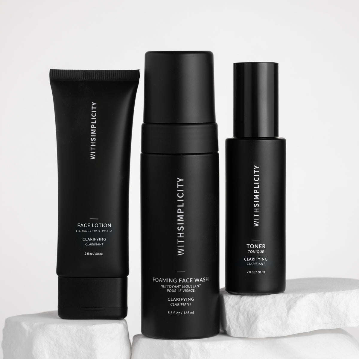 Three black skincare bottles from withSimplicity are arranged on white stone blocks against a light gray background. The products include Face Lotion, Foaming Face Wash, and Toner.