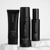 Three black skincare bottles from withSimplicity are arranged on white stone blocks against a light gray background. The products include Face Lotion, Foaming Face Wash, and Toner.