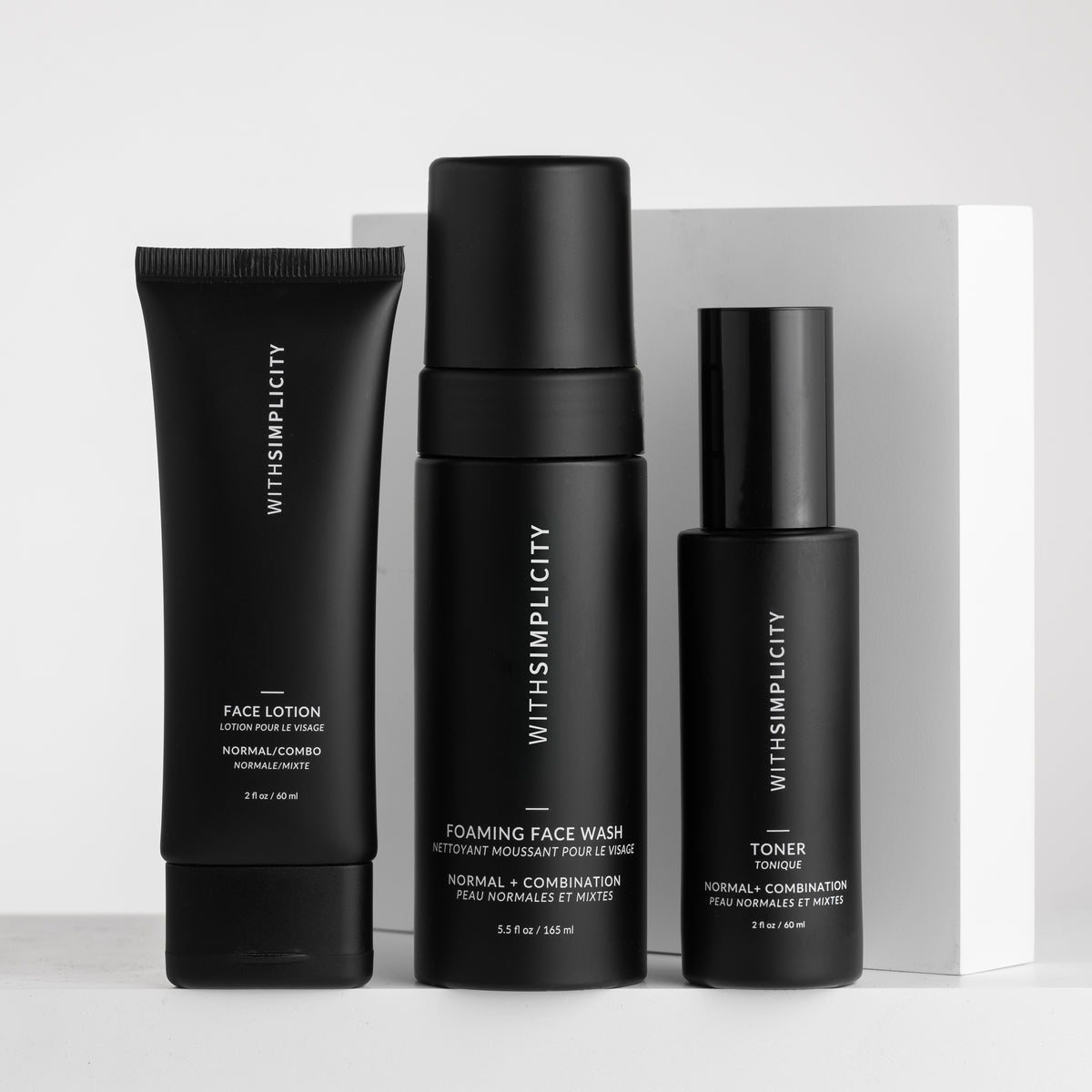 Three black skincare bottles from withSimplicity are arranged on a light gray surface against a white box background. The products include Face Lotion, Foaming Face Wash, and Toner, with white text on the labels.