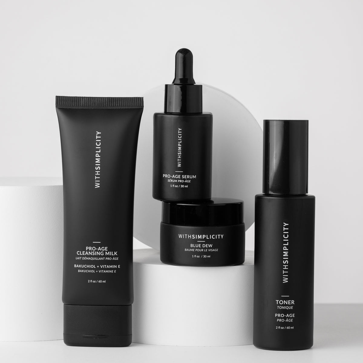 Four black skincare containers from withSimplicity arranged on white cylindrical pedestals against a light gray studio background. The products include a cleansing milk tube, a serum dropper bottle, a blue dew serum jar, and a toner bottle.