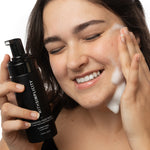 A woman with dark hair applies foaming facial wash to her face while holding a black bottle labeled withSimplicity in one hand, against a clean white background.