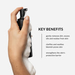 Hand holding a black foaming face wash bottle with white foam, against a light gray background. Text on image lists product benefits including: targets acne-prone skin concerns, gently removes dirt, excess oils, and residue, and finally, strengthens the skin's protective barrier