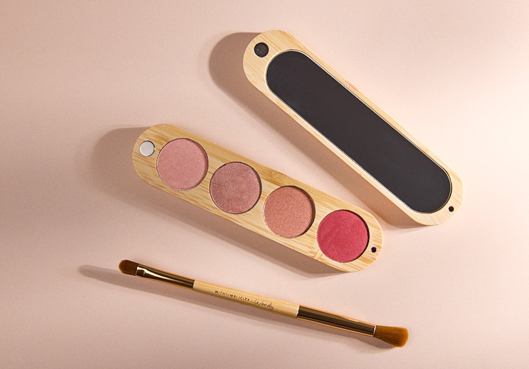 Eyeshadow palette with four pink shades, a mirror, and an eyeshadow brush on a light pink background