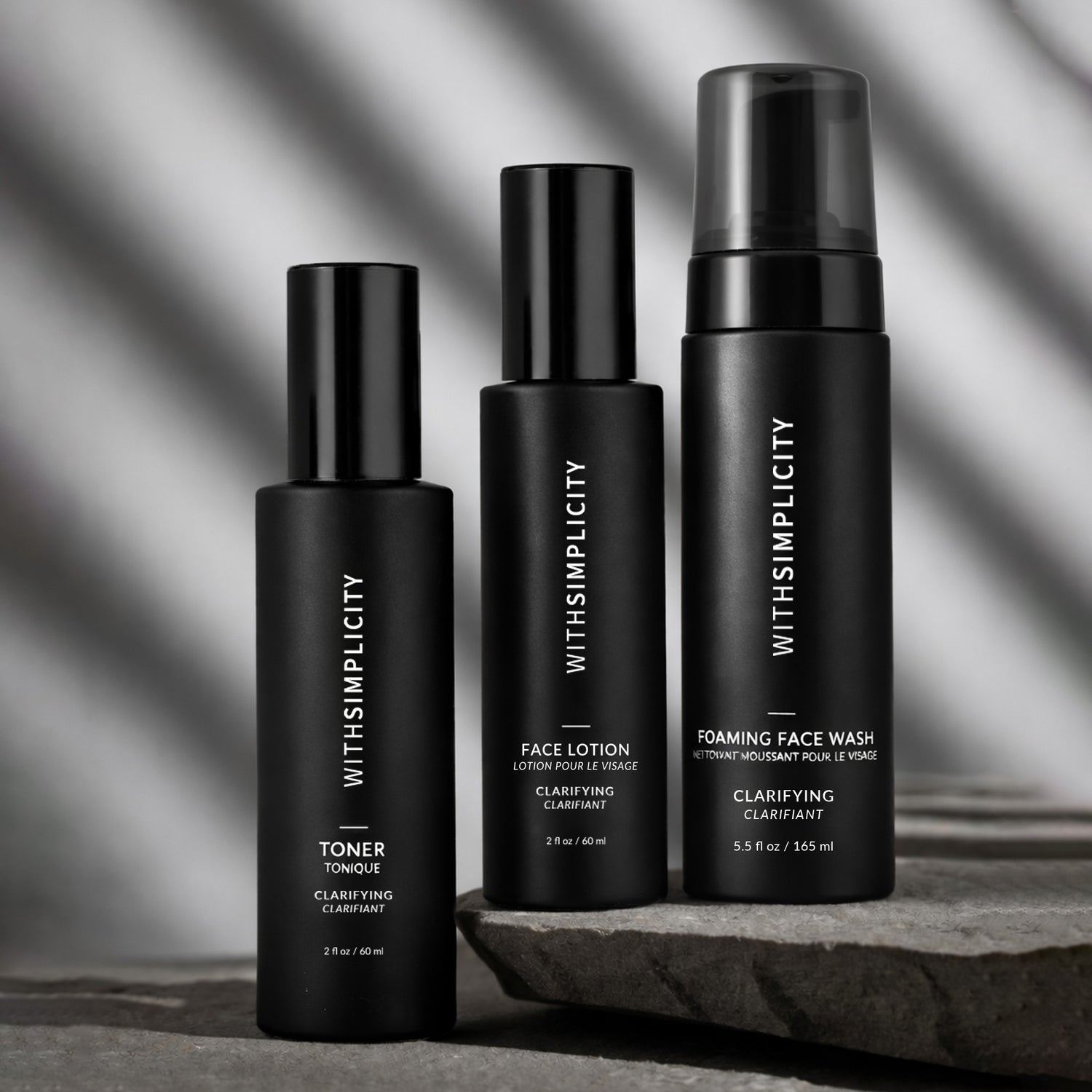 Three withSimplicity acne-prone skincare products—one toner bottle, one lotion bottle, and a foaming face wash—arranged on stone with soft striped shadows in the background.