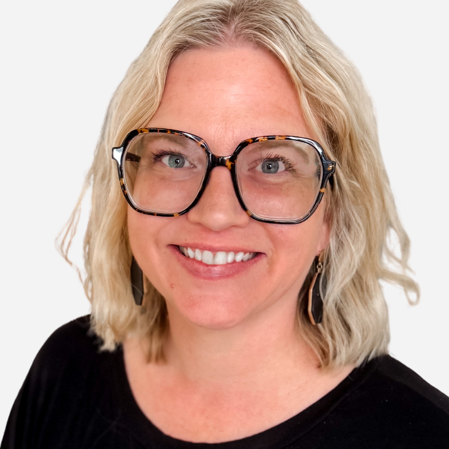 A smiling blonde woman wearing oversized square tortoiseshell eyeglasses with thick black frames and dark dangling earrings, photographed against a plain light background.