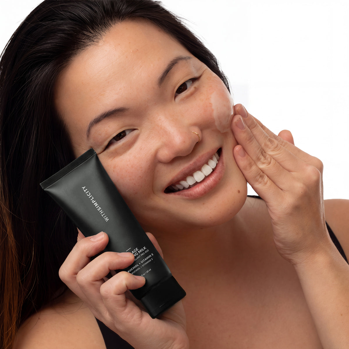 A smiling woman with dark hair applies Pro-Age Cleansing Milk her cheek while holding a black tube of withSimplicity skincare product against a clean white background.