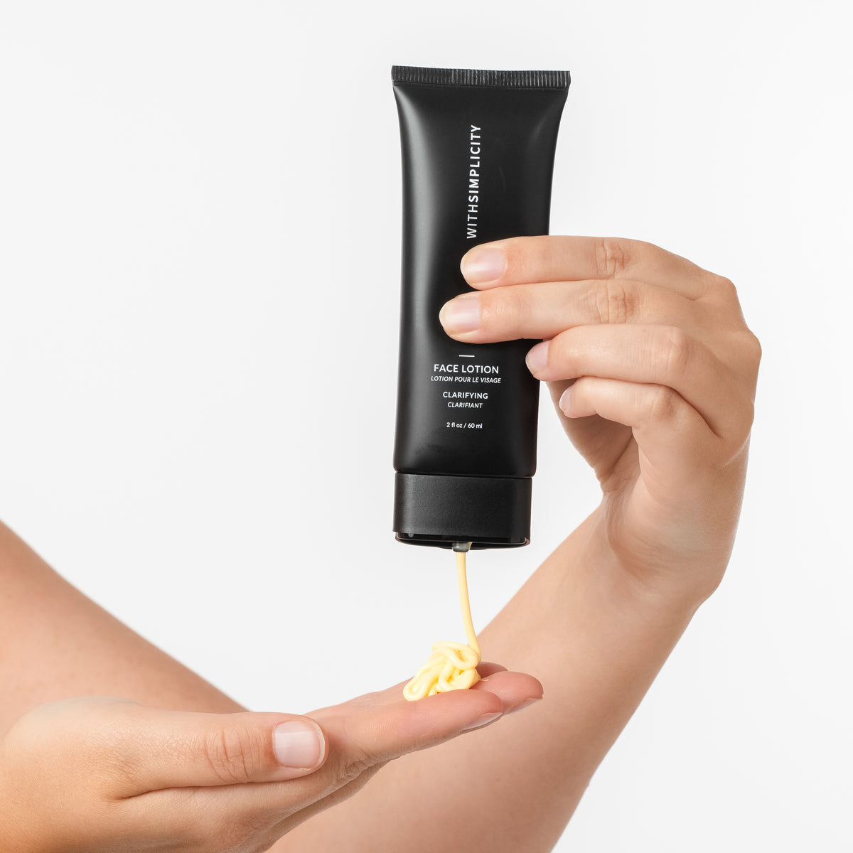 A hand holds a black squeeze tube of withSimplicity Clarifying Face Lotion, with yellow cream being squeezed onto another hand, against a clean white background.