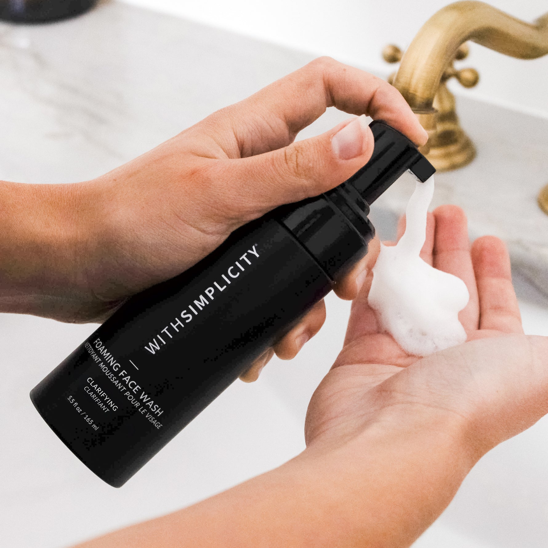 hand holding withsimplicity clarifying face wash and pumping out foam cleanser onto hand above sink
