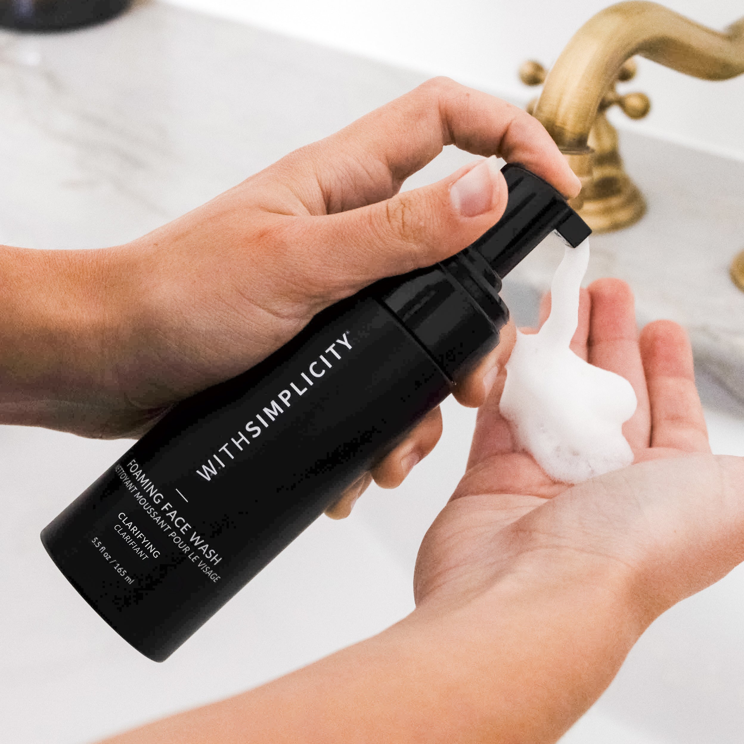 hand holding withsimplicity clarifying face wash and pumping out foam cleanser onto hand above sink