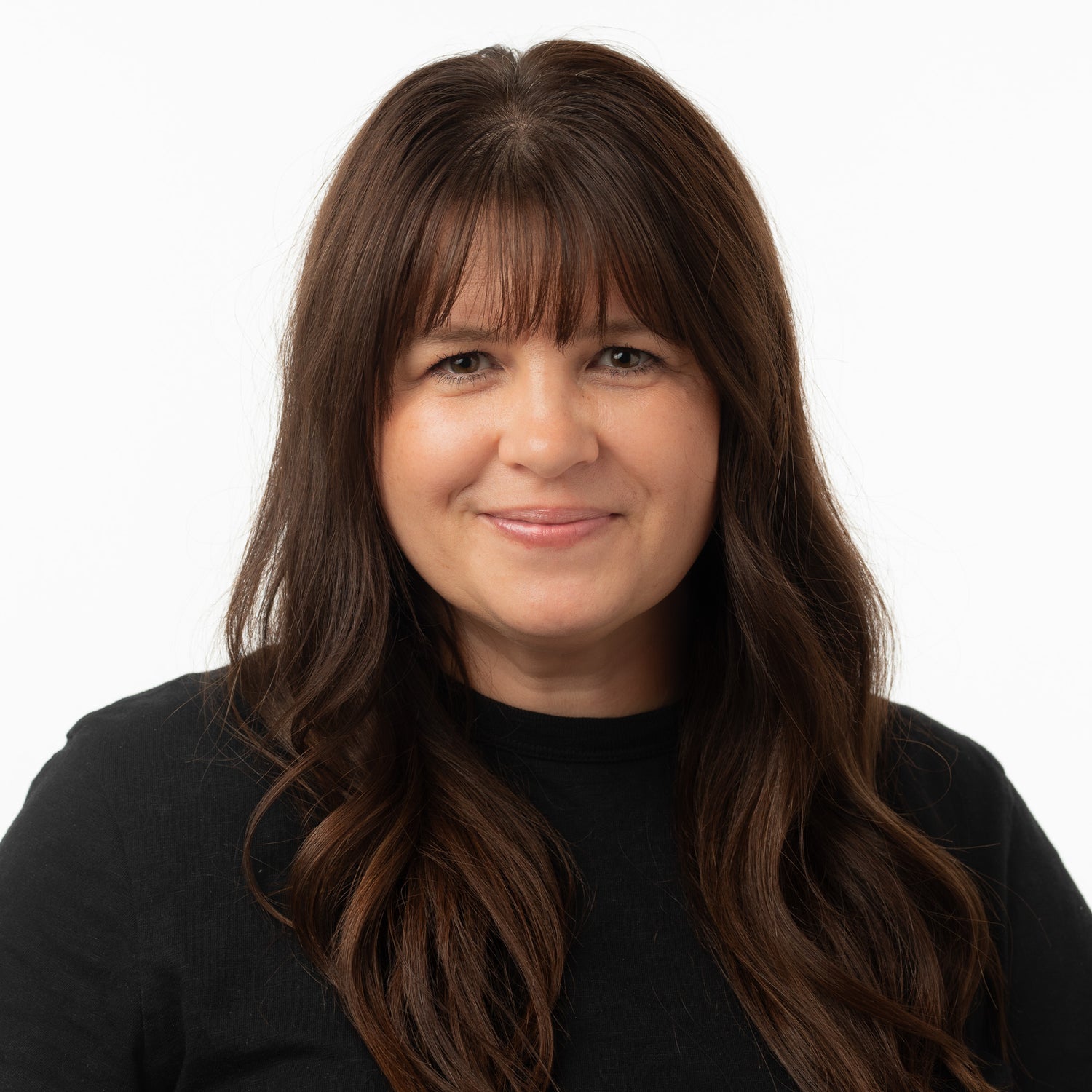 A woman with long brown hair and bangs, wearing a black top, photographed against a plain white background.