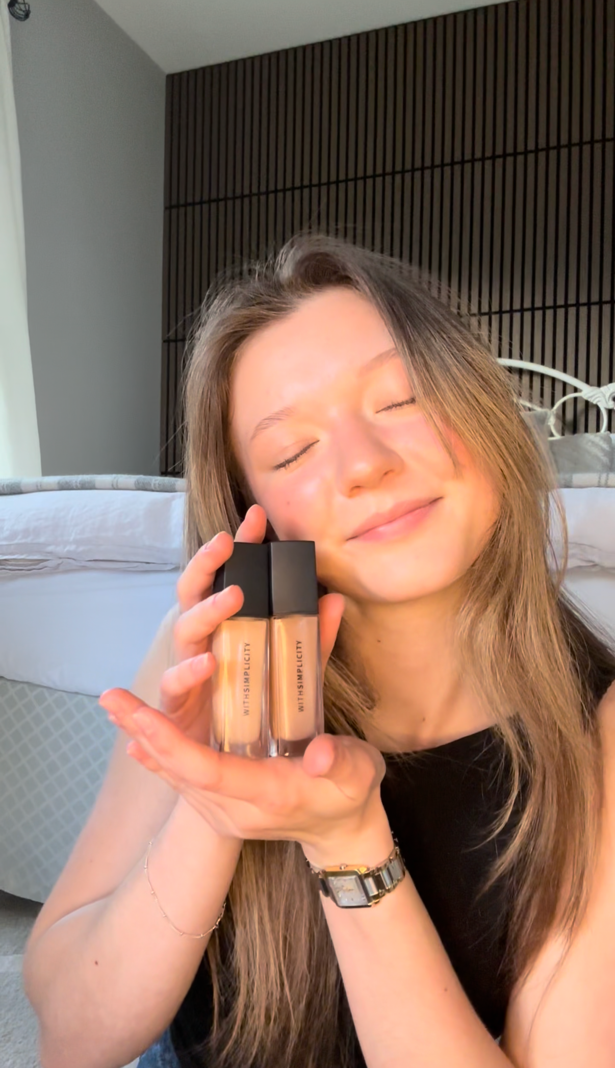 Woman holding two bottles of withSimplicity organic liquid concealer in a room with neutral decor