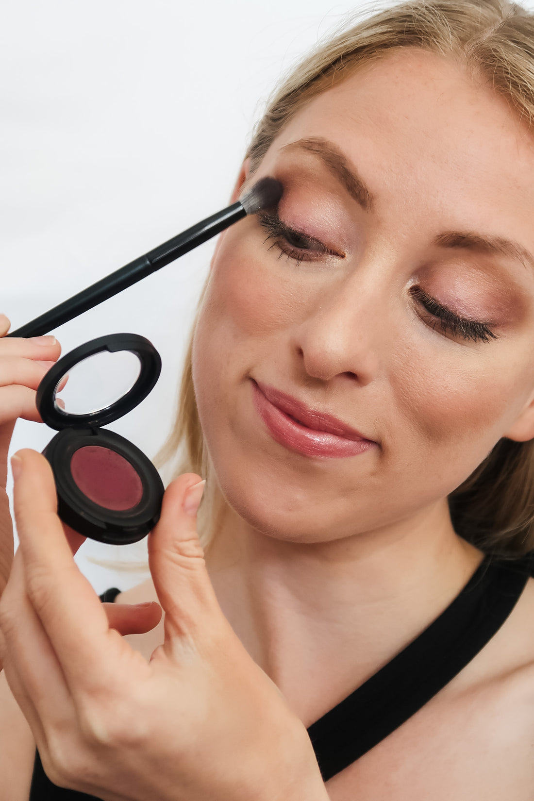 woman with blonde hair and green eyes applying makeup in a tutorial-style video.