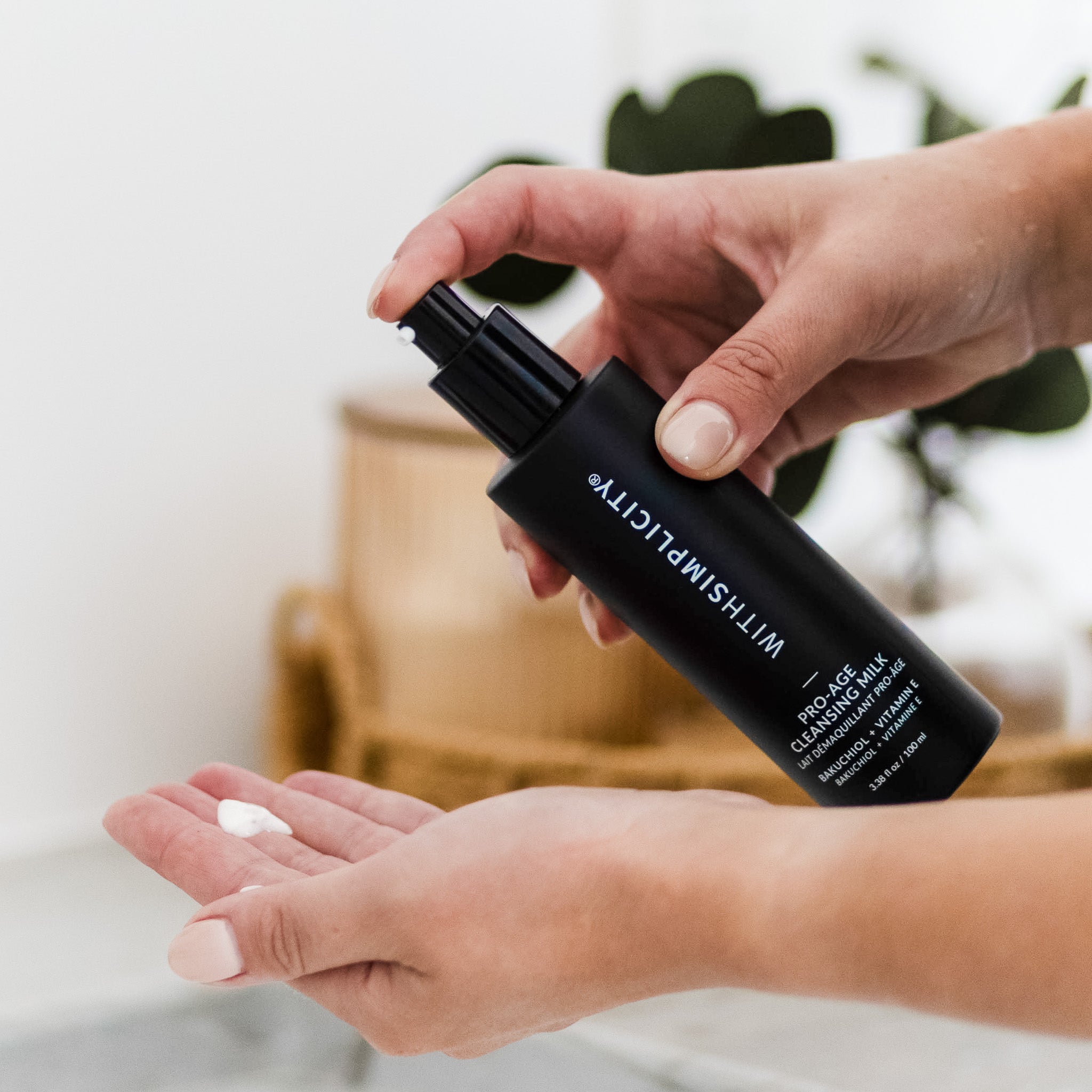 Person holding a black bottle labeled withSimplicity Pro-Age Cleansing Milk with a small amount of white cream on their palm.