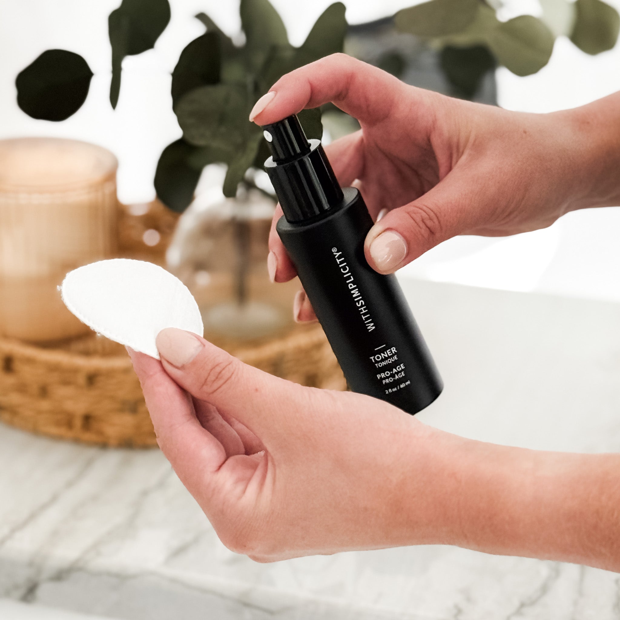 Person holding withSimplicity Pro-Age Toner with a white cotton pad, with a blurred background of plants and containers.