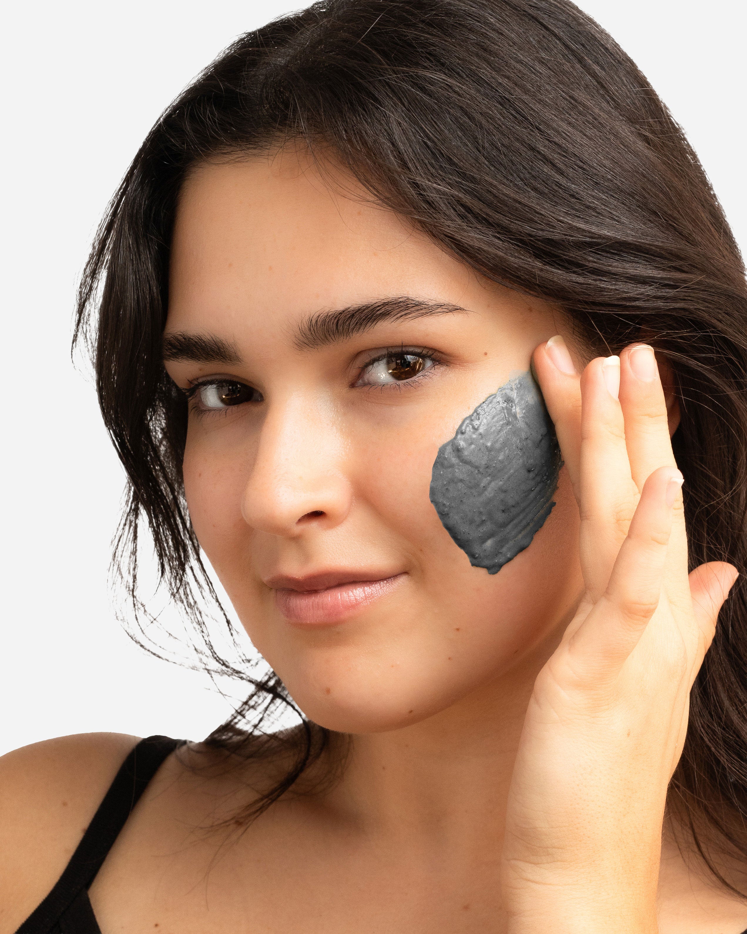 Woman applying a gray facial mask to her cheek against a neutral background