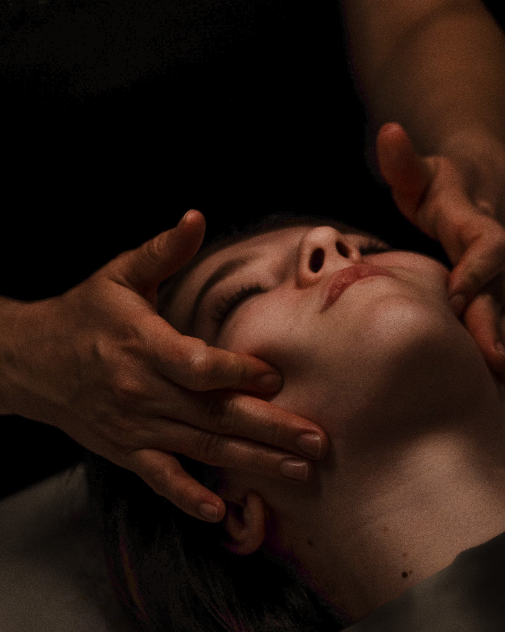 Close-up of a person's face with hands pressing down on it during a facial, in a dark setting