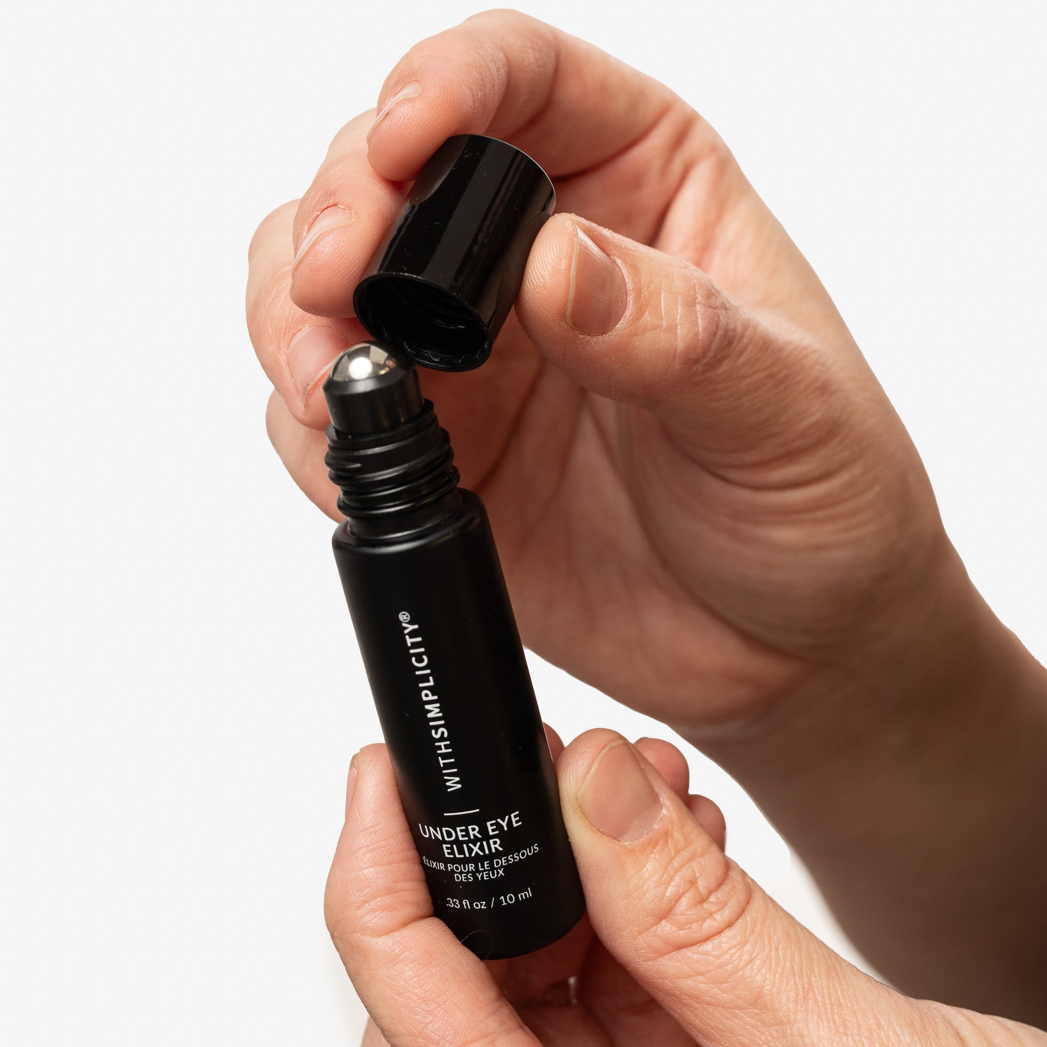 Hand holding a black rollerball bottle labeled withSimplicity Under Eye Elixir with an open cap on a white background.