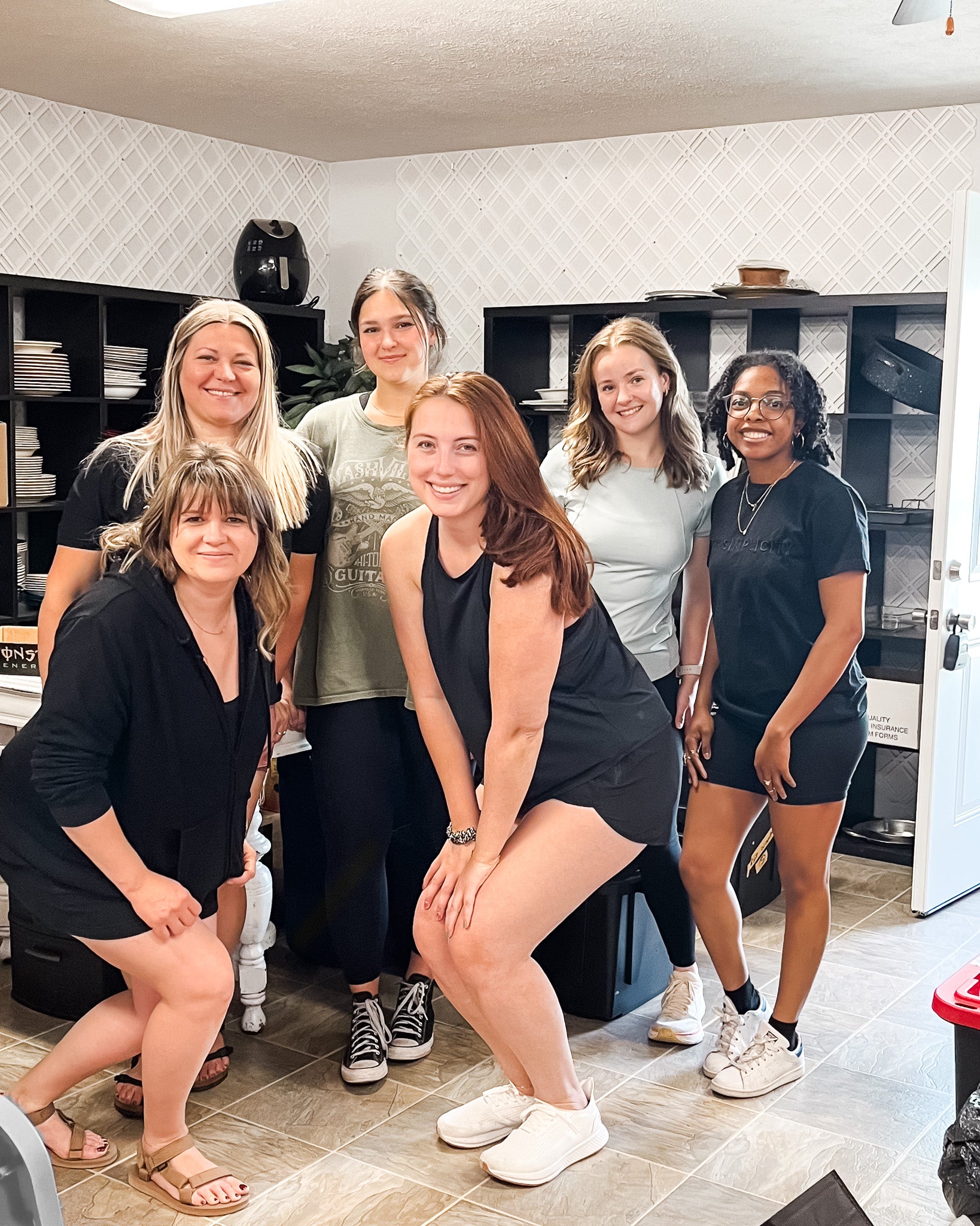 Six women posing together in a home or studio setting with shelves and tiled floors visible in the background.