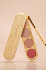 A wooden makeup palette with three round blush pans in pink, mauve, and rose tones, shown open with a matching wooden applicator brush on a soft beige background. The palette lid is engraved with the brand name 'withSimplicity x Wendy'.