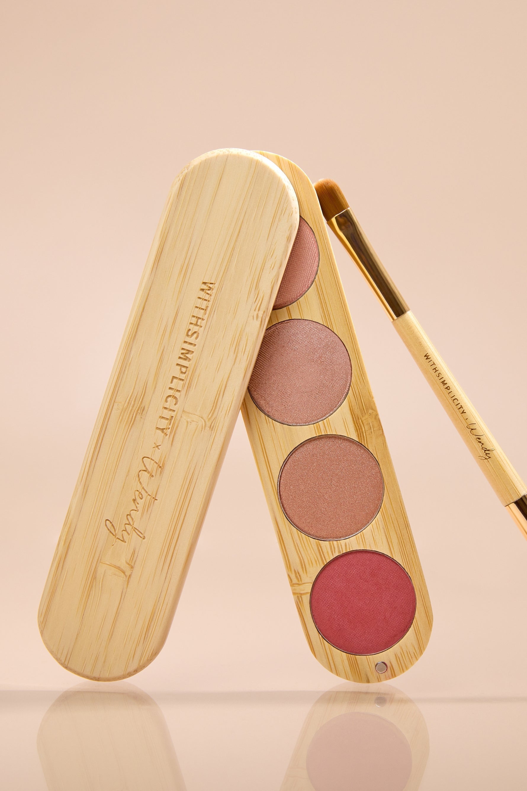 A wooden makeup palette with three round blush pans in pink, mauve, and rose tones, shown open with a matching wooden applicator brush on a soft beige background. The palette lid is engraved with the brand name 'withSimplicity x Wendy'.