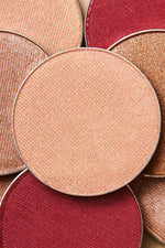 A close-up arrangement of round makeup pans in warm neutral and red shades, including a central peach-beige pressed powder surrounded by bronze, copper, and deep red tones.