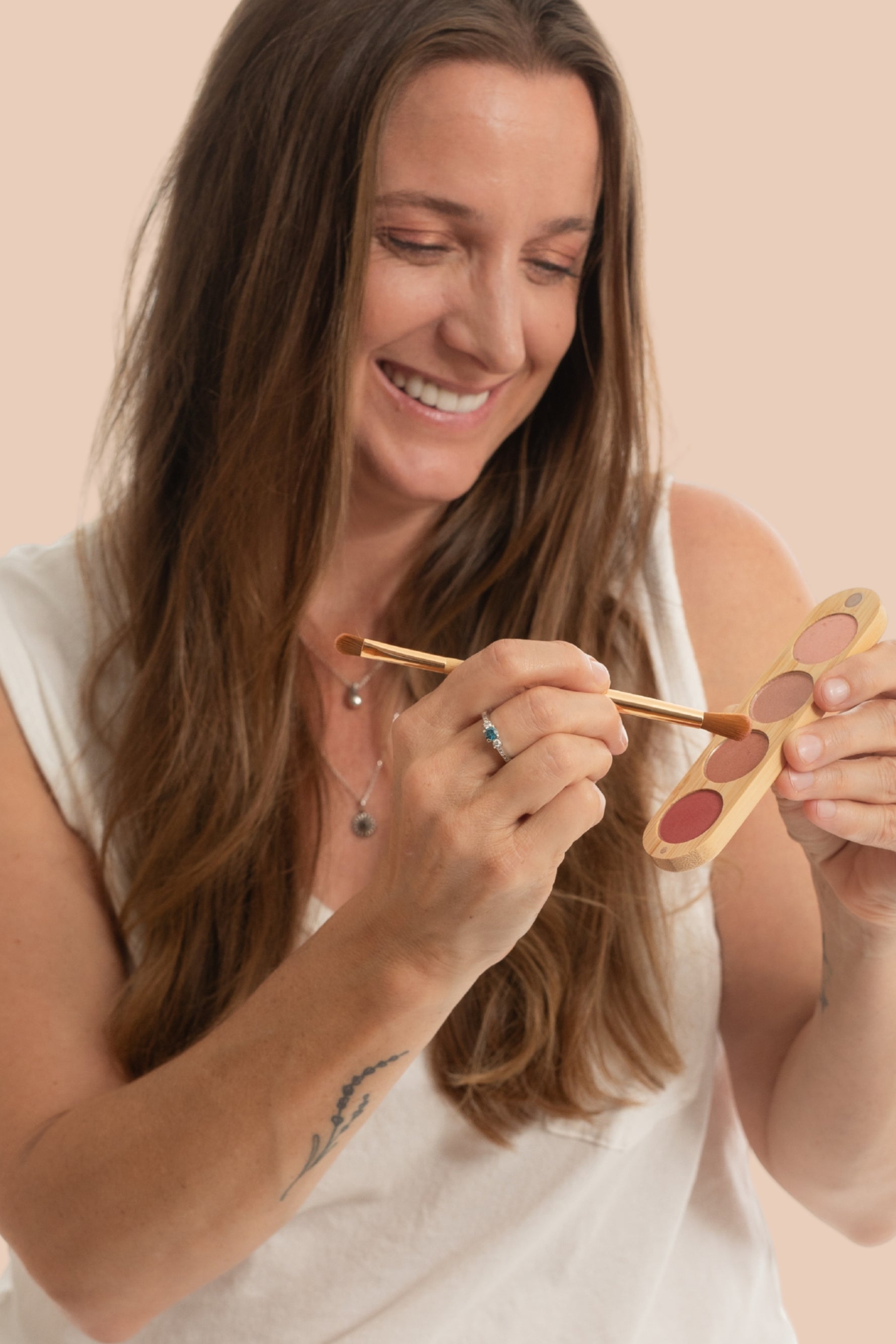 A woman with long brown hair smiles while holding a small makeup palette and applying product with a brush. She is wearing a white sleeveless top against a plain beige background.