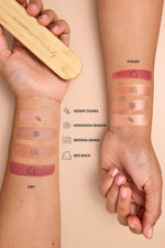 Two hands with makeup swatches on the forearms, showing different finishes and shades of a cosmetic product. A wooden eye palette is held in the left hand, and text labels identify the shades as Desert Dunes, Monsoon Season, Sedona Sands, and Red Rock
