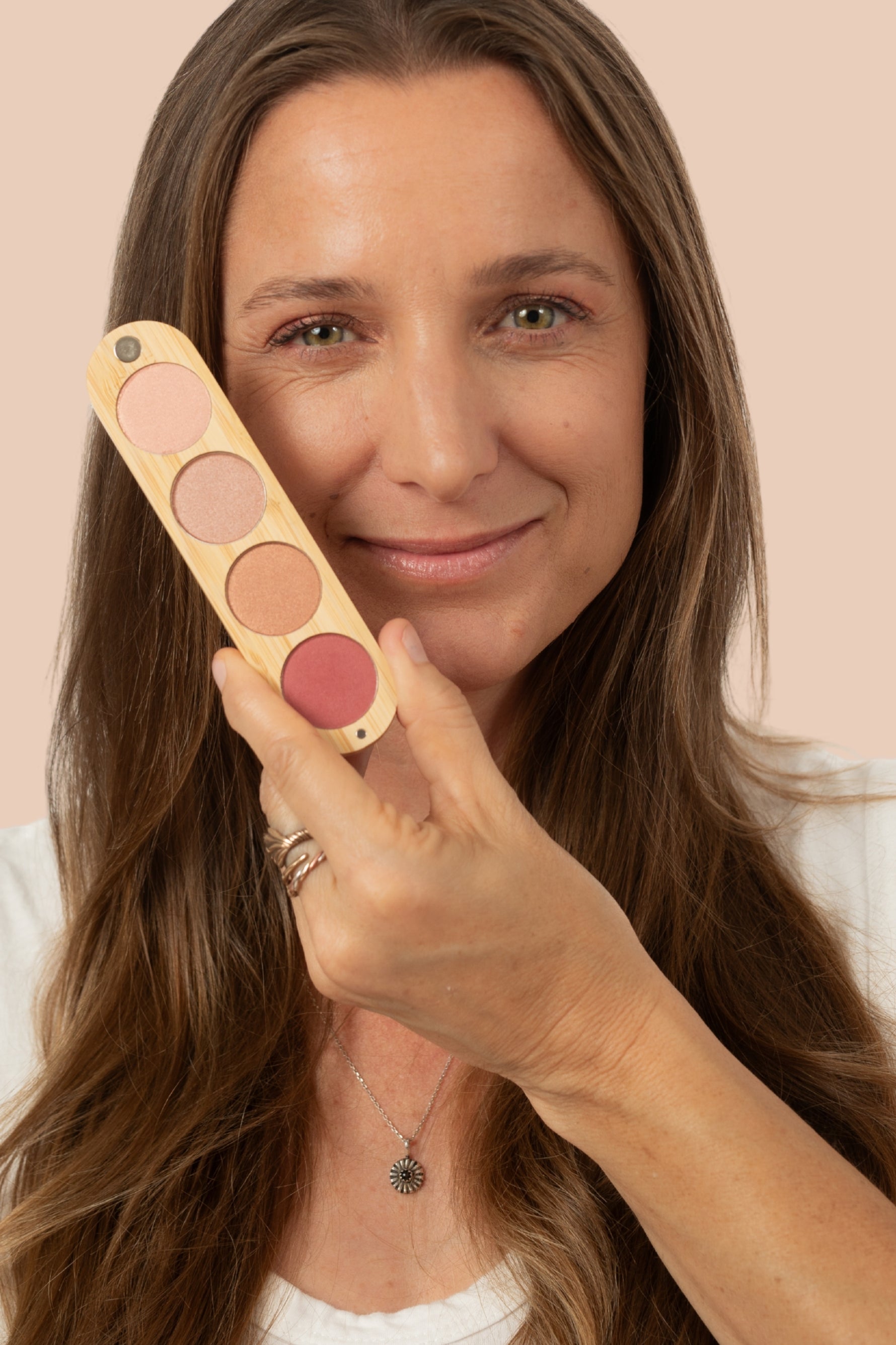A woman with long brown hair holds up a makeup palette with four round pans in pink shades against a solid pale peach background.