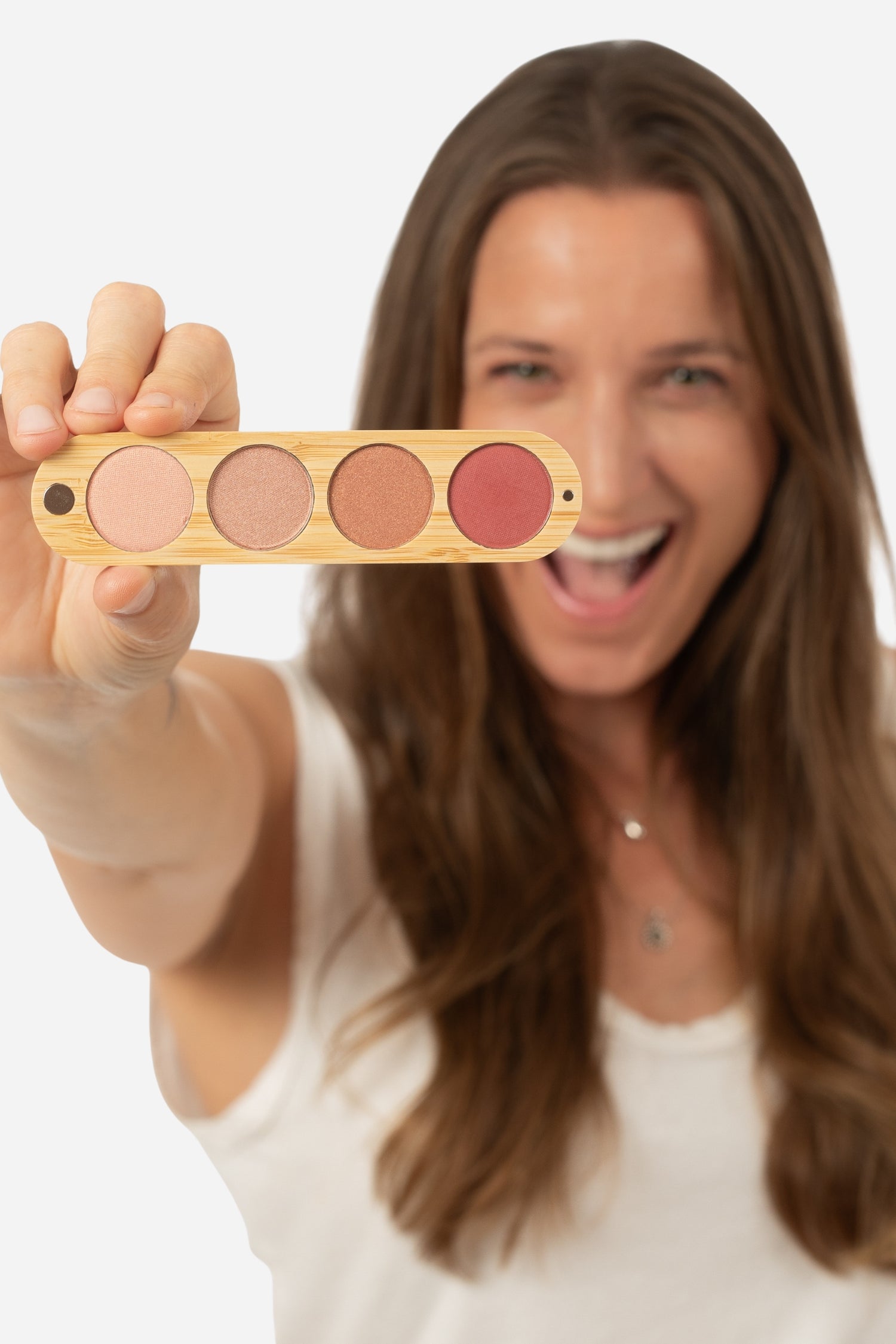 A woman with long brown hair holds up a four-pan makeup palette made of light wood. The palette contains four circular pans of makeup in neutral and rosy shades: a light beige, a medium tan, a warm brown, and a muted rose pink.