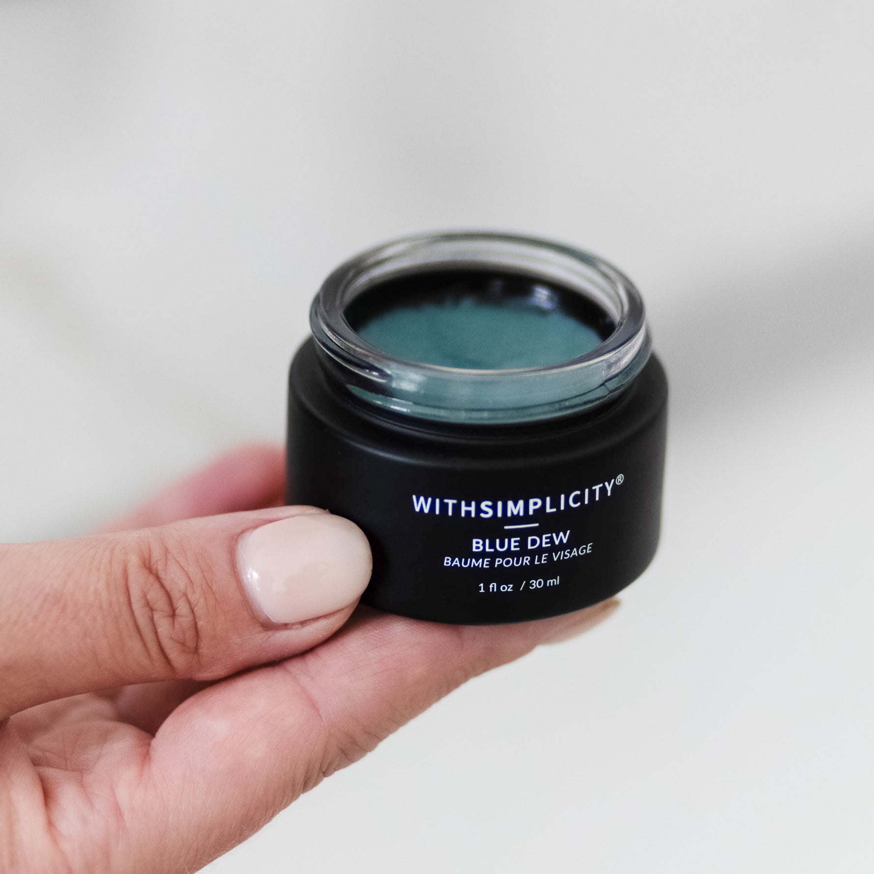 Hand holding a small jar of 'Blue Dew' cream with 'withSimplicity' branding.