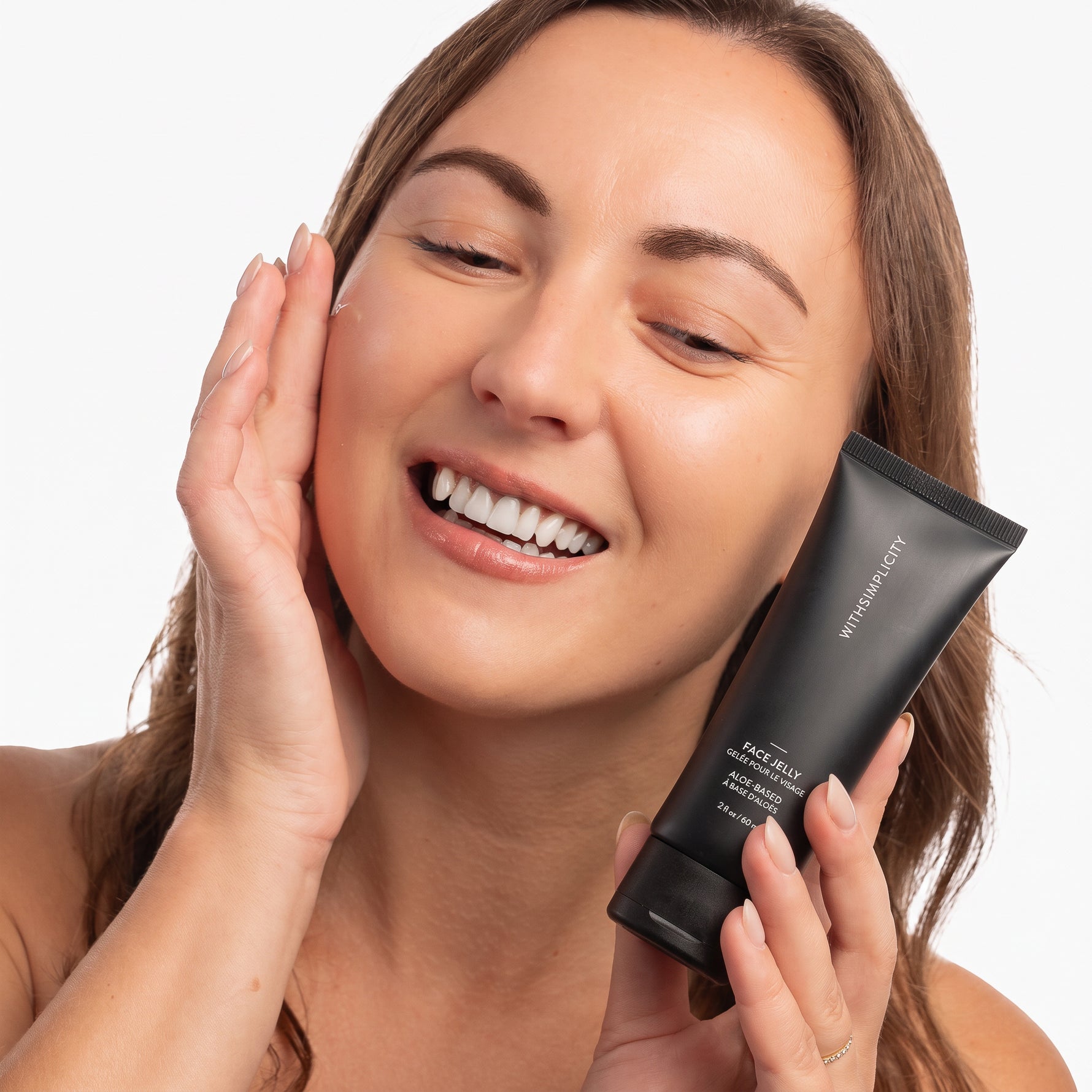 Woman applying withSimplicity Face Jelly to face and holding squeeze tube of Face Jelly in other hand against light background