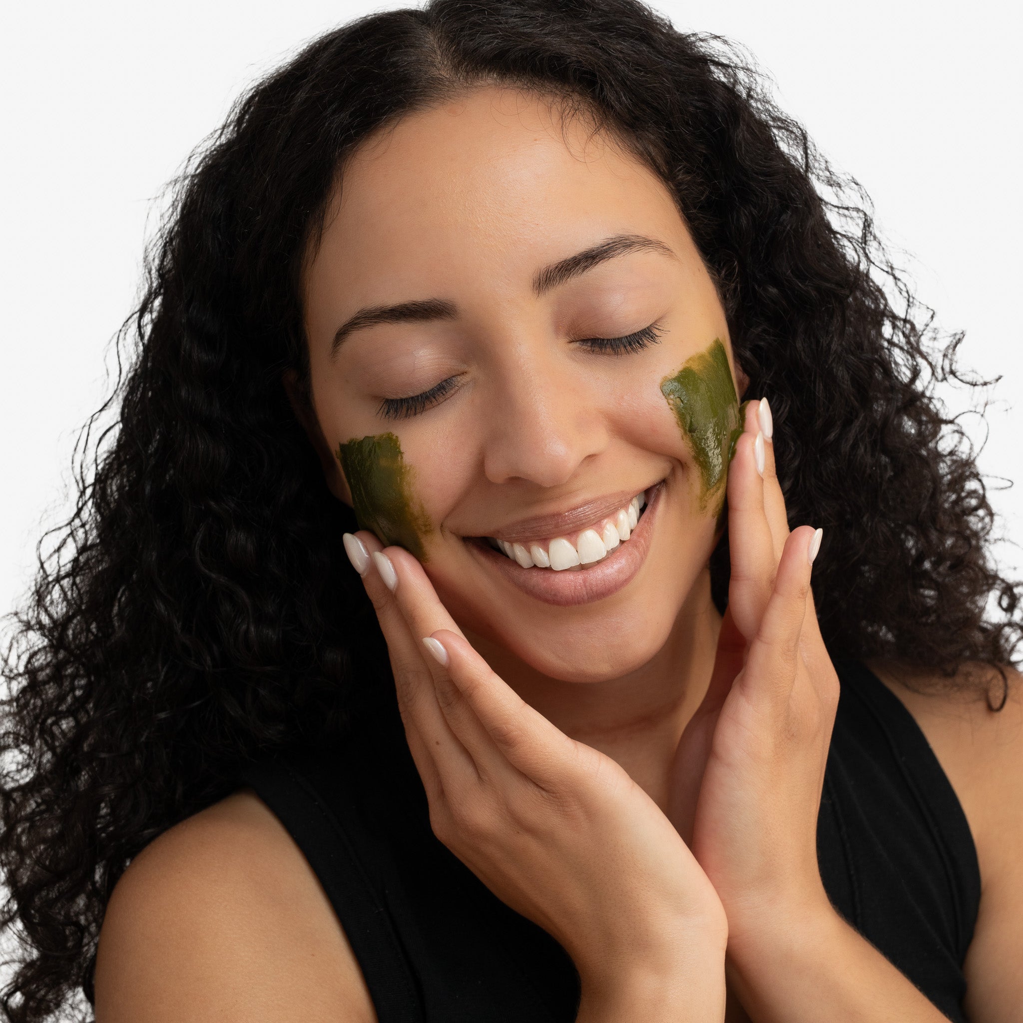 Woman applying green matcha facial masks to her cheeks with a neutral background