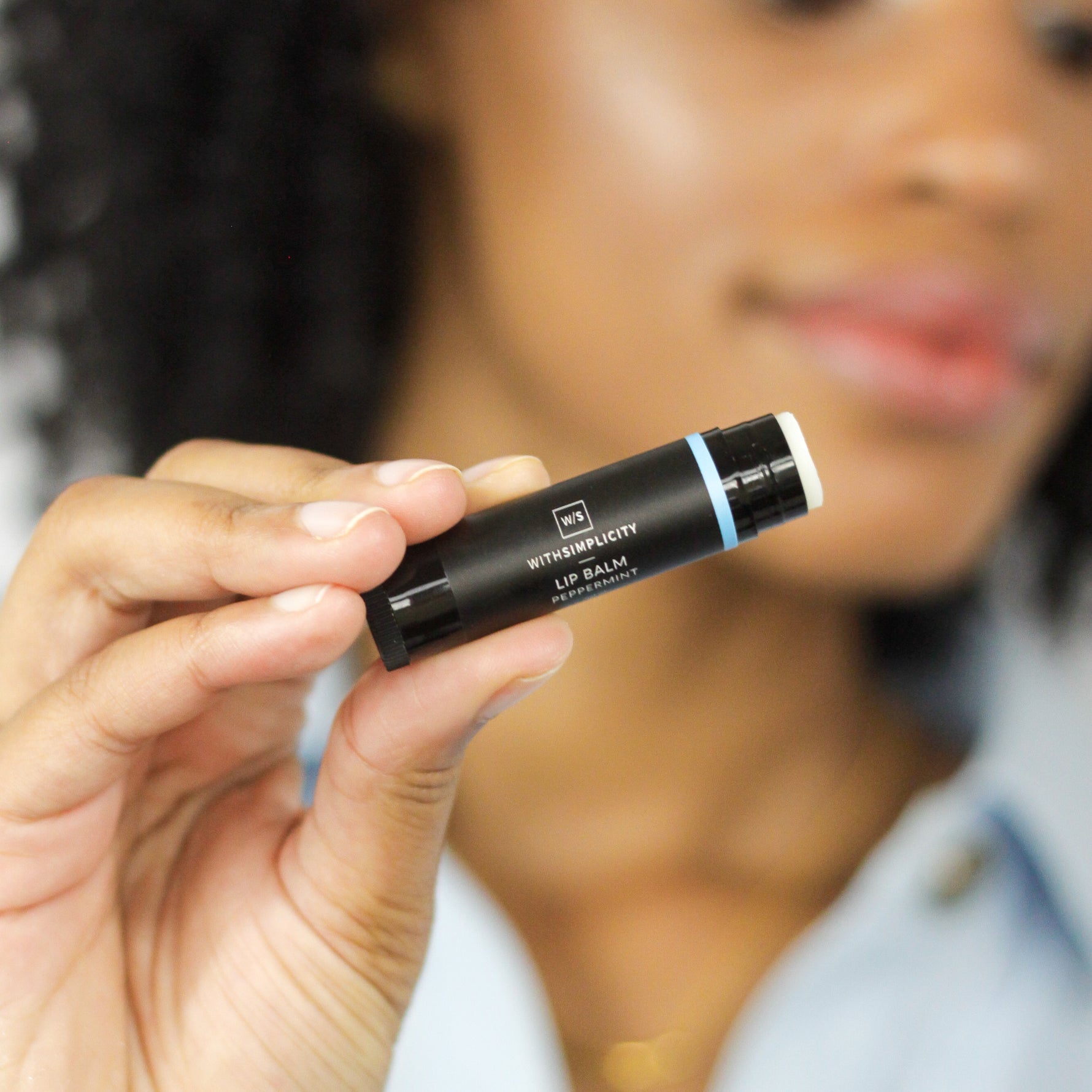 close-up of a withSimplicity Lip Balm held by a hand with a woman's face blurred in the background